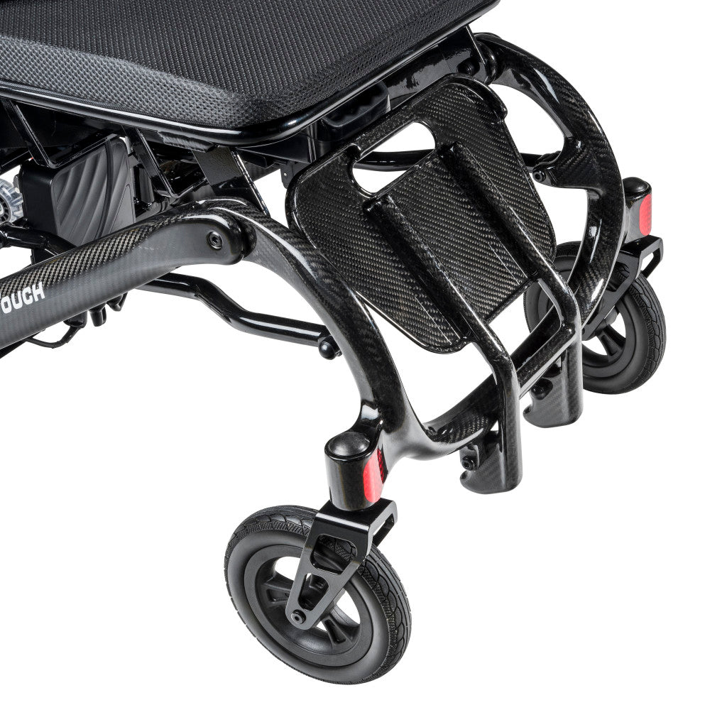 Close-up of Carbon Touch electric wheelchair front lower frame showing carbon fiber footrest platform, front swivel casters, red reflectors, and battery pack under mesh seat
