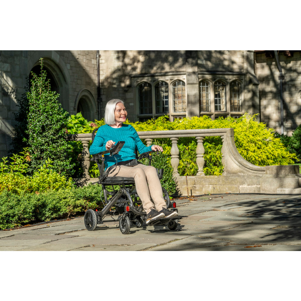 Older adult woman riding Drive Carbon Touch folding electric wheelchair outdoors on a stone pathway in a sunny garden setting, using joystick controller — available at Must in Metro Vancouver