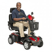 Smiling older adult man riding Drive Ventura 4-wheel mobility scooter in red and black with captain seat, dual mirrors, front basket, and all-terrain tires