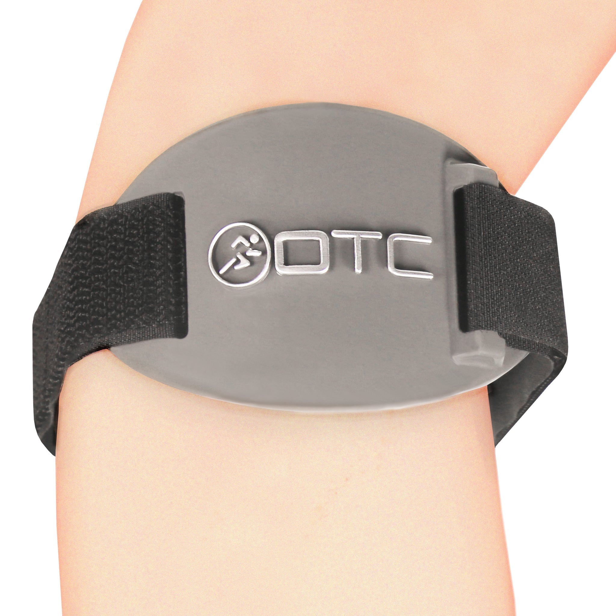 Close-up front view of OTC 2421 tennis elbow brace on forearm showing grey oval pressure pad with embossed OTC logo, black elastic strap, and velcro tabs on both sides
