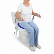 An older woman wearing a white t-shirt and light blue pants sits on a white toilet with a blue cushion, gray armrests, and a white storage bin. The photo is taken against a clean white background.